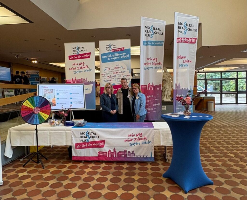 Unser Stand mit Rollups und Fahnen in der Europahalle, davor drei Lehrkräfte